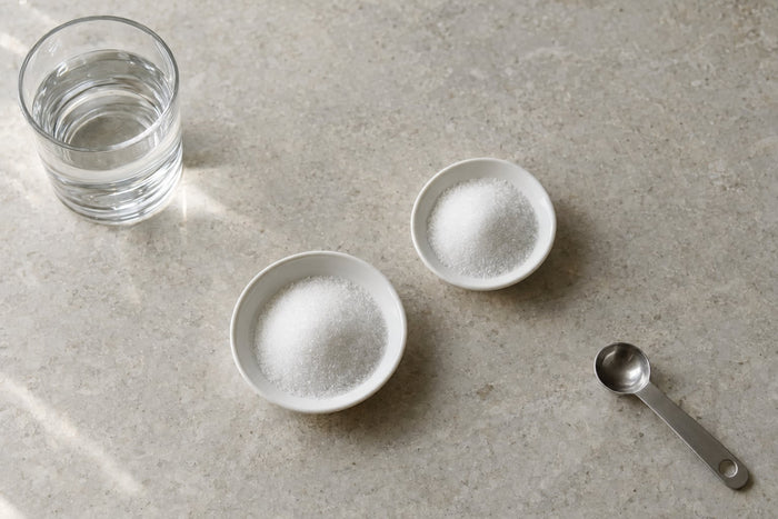 Two small white dishes of fine white crystalline powder on a pale stone counter, seen from above, with a glass of water and an empty steel measuring spoon nearby.