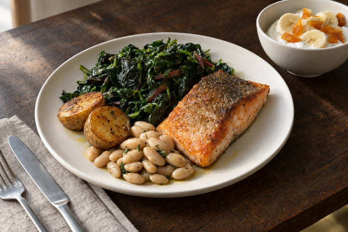 A dinner plate on a dark wooden table: seared salmon, two roasted potato halves with the skin on, white beans and wilted spinach and chard, beside a small bowl of plain yogurt topped with banana slices and chopped dried apricots.